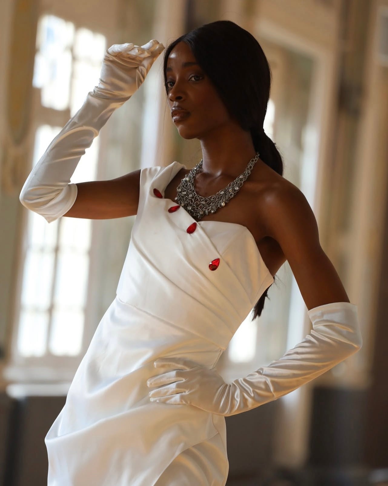 Crafted in lustrous white satin, this striking floor-length gown features an asymmetrical neckline adorned with elegant red button detailing, creating a bold yet timeless silhouette.