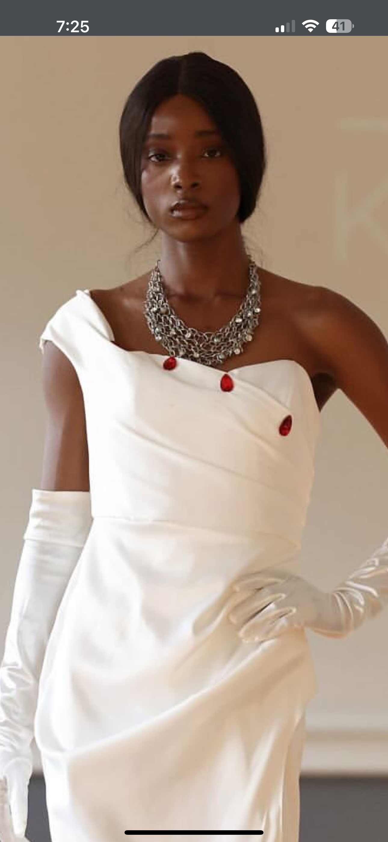 Crafted in lustrous white satin, this striking floor-length gown features an asymmetrical neckline adorned with elegant red button detailing, creating a bold yet timeless silhouette.