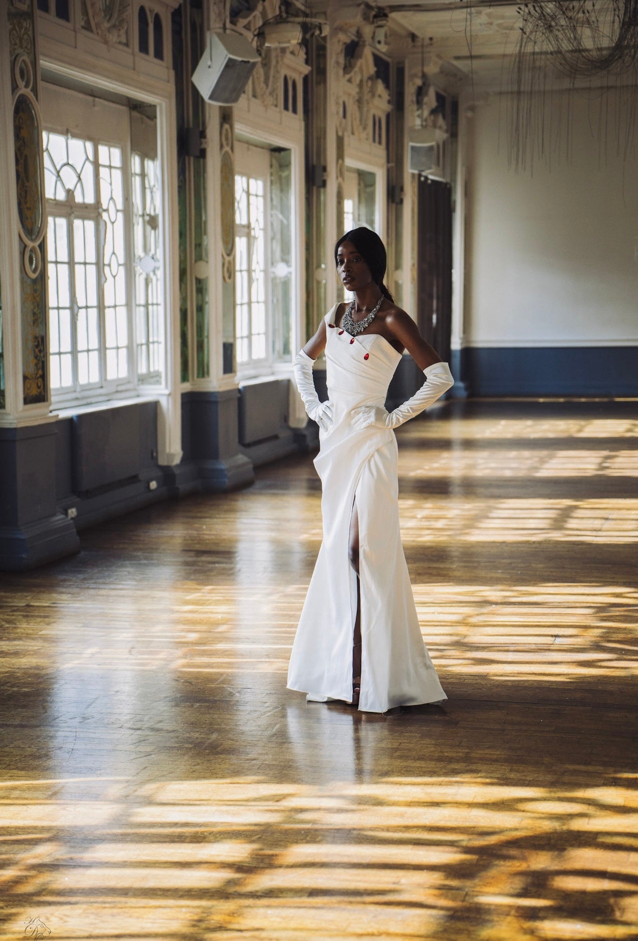 Crafted in lustrous white satin, this striking floor-length gown features an asymmetrical neckline adorned with elegant red button detailing, creating a bold yet timeless silhouette.